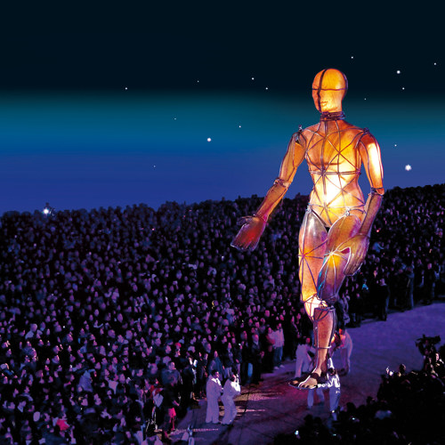 Enorm wandelend sculptuur van buizen en oranje licht loopt door menigte in het donker