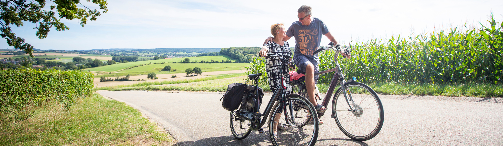 Een koppel op de fiets kijkt elkaar boven op de berg verliefd aan met achter zich een panorama over de heuvels